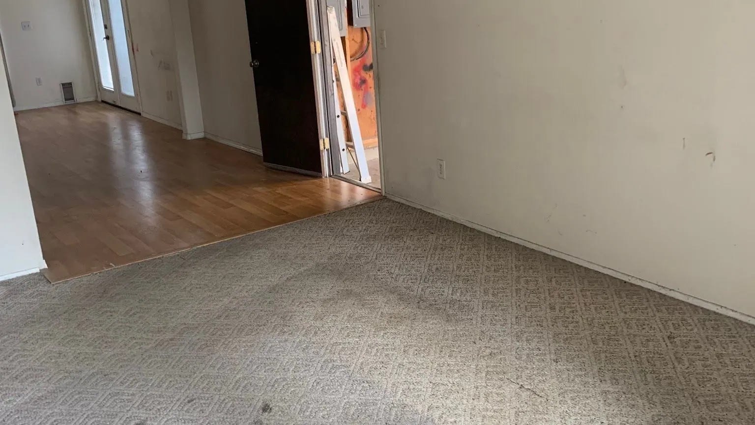 An emptied living room with patterned carpet and a laminate floor beyond