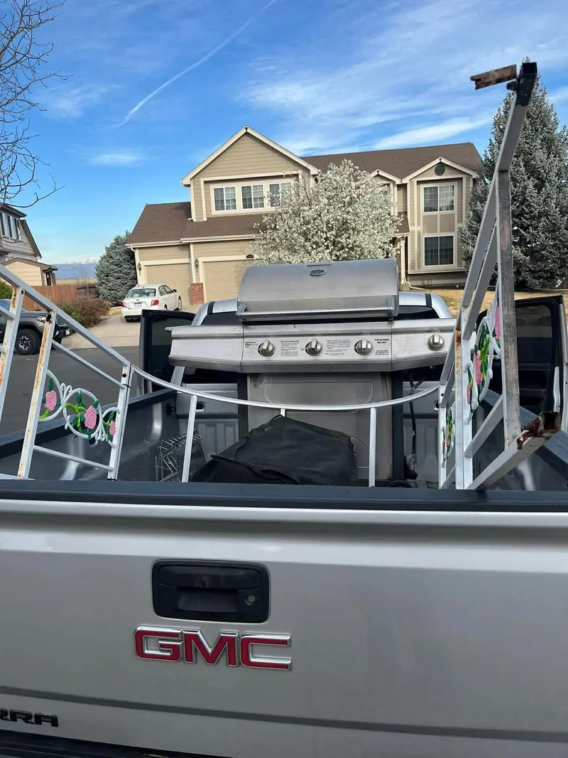 A stainless gas grill and a white metal rack in the bed of a silver GMC pickup