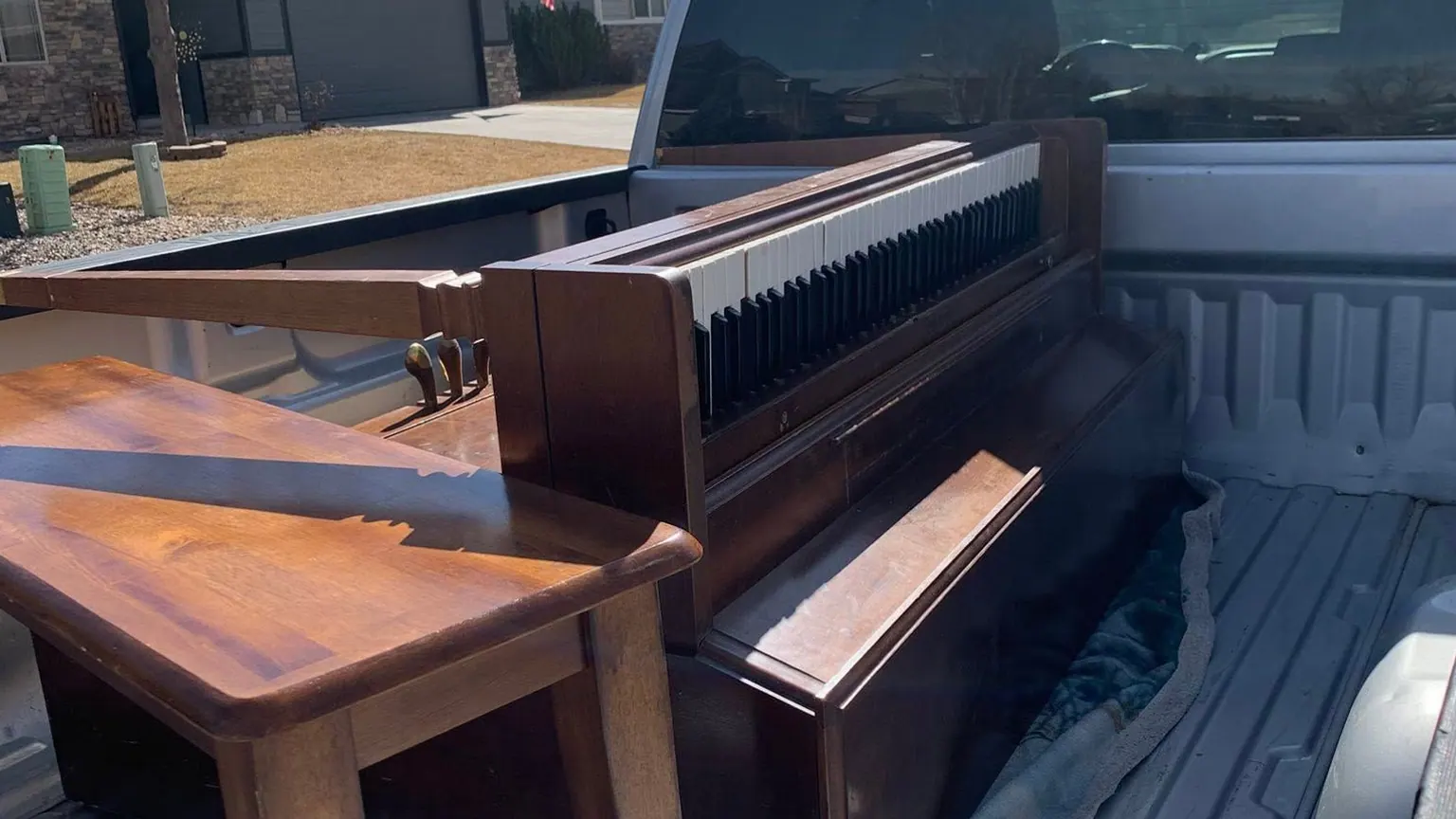 An old upright piano and a wooden side table loaded in the bed of a silver pickup