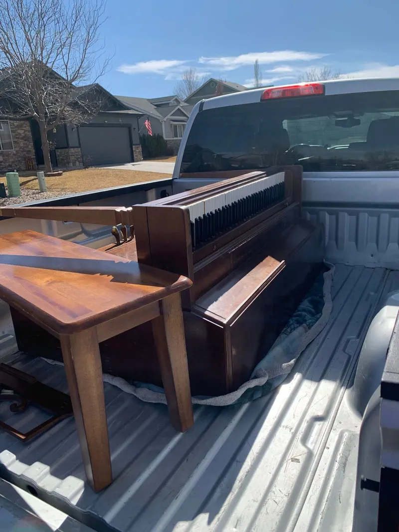An old upright piano and a wooden side table loaded in the bed of a silver pickup