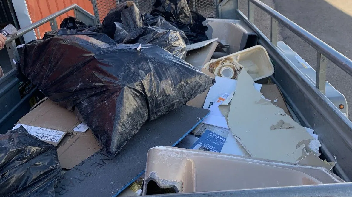 A trailer loaded with black trash bags and cardboard beside a broken sink and torn drywall