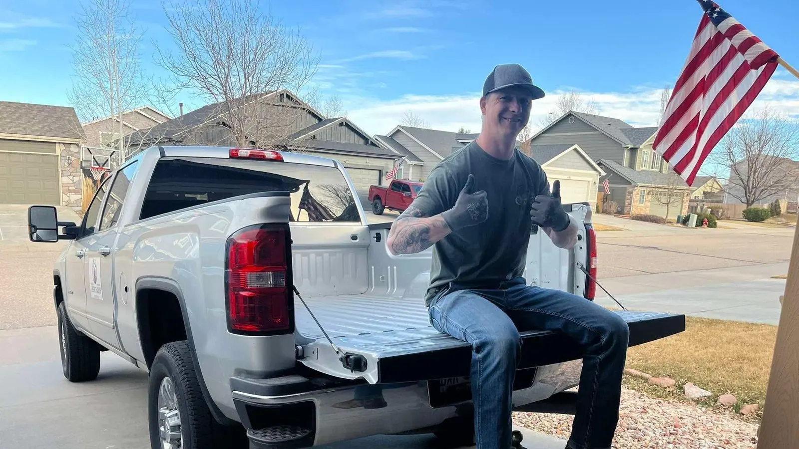 The owner sitting on the tailgate of the silver GMC pickup with an American flag in the background