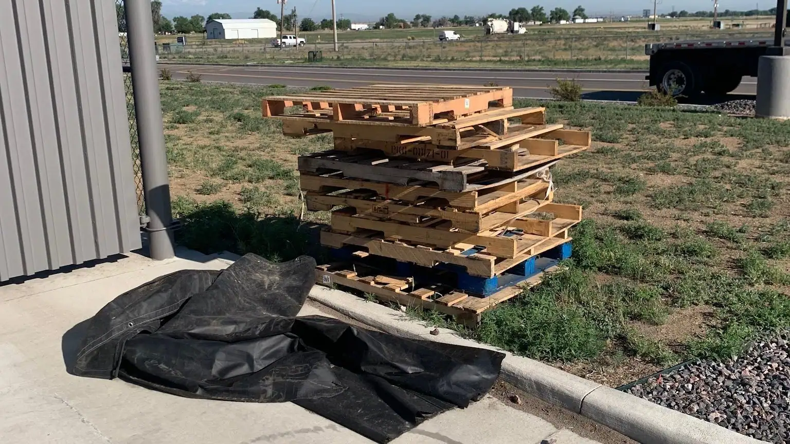 Wooden pallets stacked on the grass beside a curb, with a black tarp on the concrete