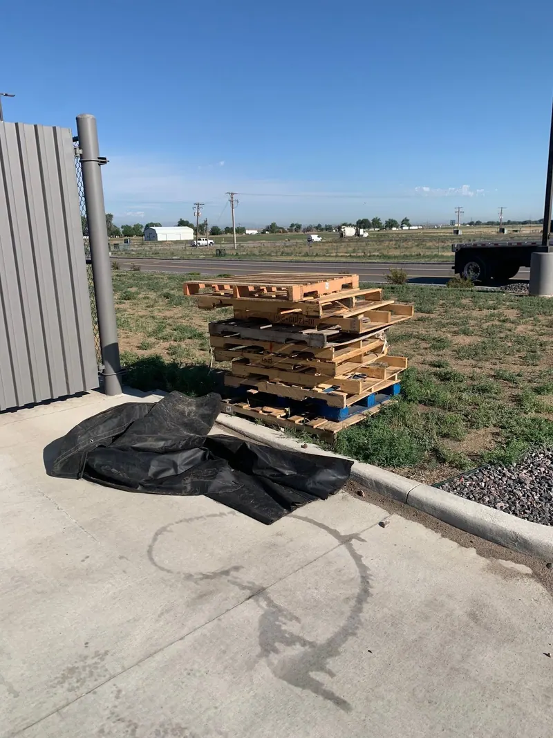Wooden pallets stacked on the grass beside a curb, with a black tarp on the concrete