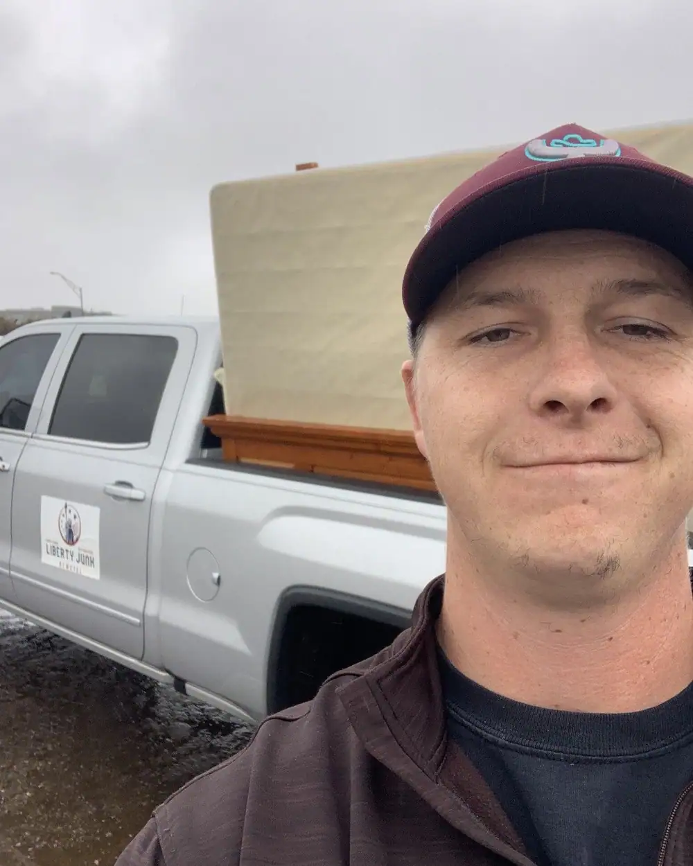 The owner in a maroon cap in front of the silver pickup with a mattress and a wooden headboard in the bed